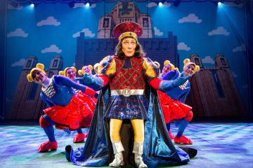 Lavalla seisoo näyttämön keskellä kuninkaalliseen pukuun pukeutunut esiintyjä, jota reunustavat tanssijat kirkkaan punaisissa ja sinisissä asuissa, joissa on karhunkorvakorvapanta. Linnan tausta ja dramaattinen valaistus luovat eloisan teatteritunnelman.