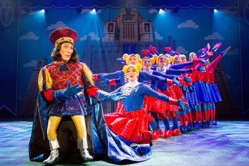 Lavalla esiintyy mies värikkäässä kuninkaallisessa puvussa ja korkeassa hatussa edessä, ja kuorossa on esiintyjiä kirkkaissa sinisissä, punaisissa ja keltaisissa puvuissa, jotka poseeraavat dramaattisesti lavan valojen alla.