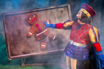 Värikkääseen keskiaikaiseen pukuun pukeutunut henkilö osoittaa dramaattisesti tarjottimella olevaa suurta piparkakkumieskeksiä, jota ympäröi teatterisavu lavalla.