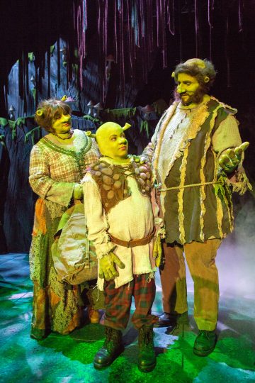Kolme värikkäisiin örkkiasuihin pukeutunutta näyttelijää seisoo lavalla vihreän valaistuksen ja metsämaisemien ympäröimänä ja esittää Shrek-musikaalin hahmoja. Keskellä olevalla hahmolla on vihreä kaljupäähine ja ruskea liivi.