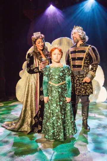 Kolme keskiaikaiseen asuun pukeutunutta henkilöä seisoo lavalla valonheittimien alla. Edessä on vihreä kukkamekkoon pukeutunut nuori tyttö, jota reunustavat nainen ja mies, joilla molemmilla on kruunut ja koristeelliset kaavut, mikä viittaa kuninkaalliseen perhekohtaukseen.