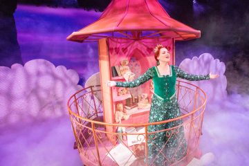 Vihreään mekkoon ja kruunuun pukeutunut nainen laulaa iloisesti kädet ojennettuina vaaleanpunaisella parvekkeella, joka on koristeltu kirjoilla ja nallekarhuilla.