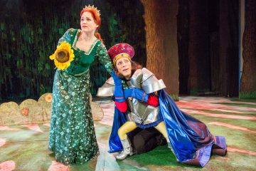 Vihreään pukuun ja kruunuun pukeutunut nainen pitelee auringonkukkaa, kun taas kiiltävään haarniskaan ja värikkääseen viittaan pukeutunut henkilö polvistuu kädestä pitäen lavalla, jonka taustalla on metsä.