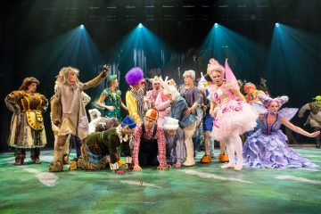 Ryhmä näyttelijöitä värikkäissä, omituisissa eläin- ja satuasuissa esiintyy lavalla, jossa on siniset valonheittimet ja vihreä lattia, mikä viittaa vilkkaaseen teatteriesitykseen.