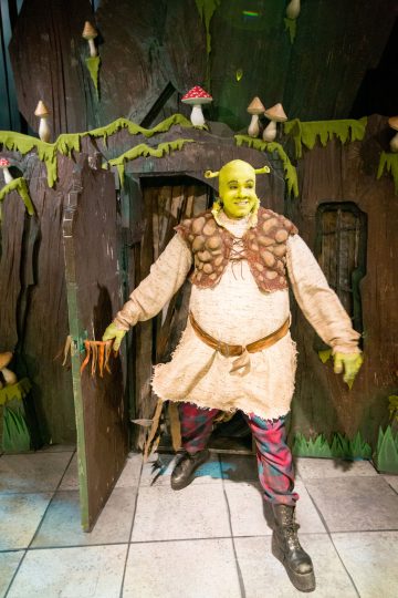 Shrekiksi pukeutunut henkilö, jolla on vihreä örkkiasu ja ruskea liivi, astuu ulos puisesta ovesta, jossa on sieniä ja viherkasveja, jotka muistuttavat Shrekin animaatioelokuvasta tuttua suomajaa.