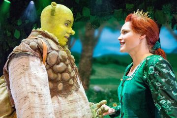Kaksi näyttelijää lavalla puvuissa: toinen on Shrek, jolla on vihreä iho ja örkki-piirteet, ja toinen on Fiona, jolla on punaiset hiukset ja vihreä mekko, ja he seisovat vastakkain metsätaustaa vasten.