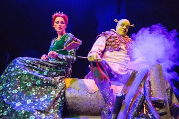 Kaksi pukumiestä istuu nuotion äärellä lavalla; toinen on pukeutunut prinsessa Fionaksi vihreään mekkoon ja kruunuun, toinen Shrekiksi vihreällä örkkimäki-meikillä ja korvilla, ja hän pitää tikkua nuotion yllä, josta nousee violettia savua.