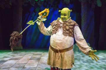 Shrekiksi pukeutunut henkilö, jolla on vihreä kasvomaali, korvien proteesit ja keskiaikaiset vaatteet, seisoo lavalla ja pitää kädessään suurta auringonkukkaa. Taustalla on puita ja sininen valaistus.