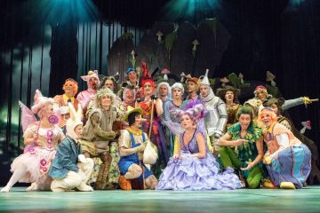 Suuri joukko värikkäisiin, omituisiin pukuihin pukeutuneita näyttelijöitä poseeraa yhdessä lavalla satuhahmoja ja fantasiahahmoja muistuttaen, ja heidän takanaan on metsäaiheinen tausta.