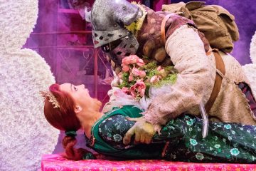 Vihreään mekkoon ja kruunuun pukeutunut nainen makaa vaaleanpunaisella alustalla ja hymyilee ritarikypärään ja -asuun pukeutuneelle henkilölle, joka kumartuu hänen ylitseen pitäen kädessään vaaleanpunaista kukkakimppua; taustalla oudot näyttämöasetelmat.