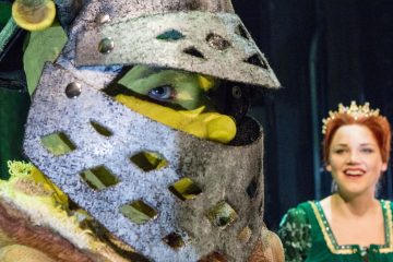 Etualalla seisoo yksityiskohtaiseen jättiläispukuun ja kypärään pukeutunut henkilö, jonka ainoa silmä on näkyvissä. Taustalla hymyilee vihreään mekkoon ja kruunuun pukeutunut nainen, jolla on punaiset hiukset. Molemmat näyttävät olevan lavalla.