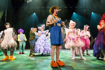 Lavalla seisoo joukko esiintyjiä värikkäissä, omituisissa puvuissa. Keskushahmo on pukeutunut Pinokkio-pukuun, jolla on pitkä nenä ja olkihattu, ja häntä ympäröivät muut sadusta inspiroituneisiin asuihin pukeutuneet henkilöt. Näyttämön valot valaisevat kohtausta.