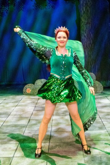 Prinsessaksi pukeutunut nainen poseeraa lavalla vihreään pukuun ja kruunuun pukeutuneena, ja pitää osaa hulmuavasta viitastaan ylhäällä metsäaiheista taustaa vasten.