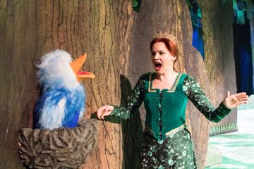 Nainen, jolla on vihreä keskiaikainen mekko ja kruunu, seisoo lavalla, laulaa kädet ojennettuina ja vastassaan on sinivalkoinen lintunukke pesässä, joka on kiinnitetty suureen puuhun.