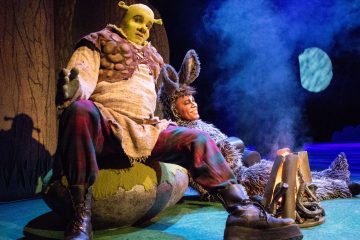 Kaksi Shrekiksi ja aasiksi pukeutunutta näyttelijää istuu lavalla leirinuotion äärellä, ja sininen näyttämövalaistus sekä puu- ja kuutausta toistavat Shrek-musikaalin kohtauksen.