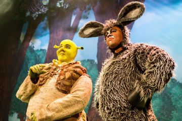 Lavalla esiintyy kaksi pukumiestä, joista toinen on pukeutunut Shrekiksi, jolla on vihreä iho ja örkinkorvat, ja toinen Aasiksi, jolla on karvainen harmaa puku ja pitkät korvat. Taustalla näkyy metsäkuva.