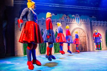 Kuusi esiintyjää värikkäissä, kiiltävissä puvuissa, vaaleissa peruukissa ja punaisissa kengissä seisoo lavalla keskiaikaisen linnan taustan edessä kirkkaassa teatterivalaistuksessa.