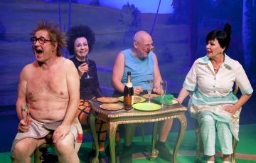 Neljä aikuista istuu pienen pöydän ääressä juomien ja ruoan kanssa nauraen ja keskustellen. Yksi mies on ilman paitaa ja silmälaseja, toisella on toppi, teatterimeikattu nainen pitelee viinilasia, ja toisella naisella on ruudullinen esiliina.