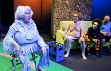 Valkoiset kiharat hiukset omaava esiintyjä, jolla on sininen röyhelöasu, istuu kirkkaasti valaistulla näyttämöllä sohvalla kolmea ihmistä vastapäätä, joista yksi on lähes alaston vanhempi mies, toinen mustaan ja oranssiin pukuun pukeutunut henkilö ja kolmas kalju mies.