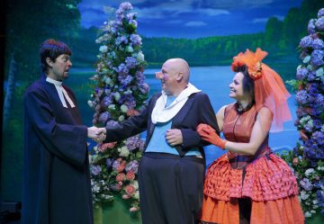 Lavalla esiintyy kolme värikkäisiin pukuihin pukeutunutta näyttelijää, joista toinen on pukeutunut papiksi ja kättelee kaljua miestä juhlapuvussa. Heidän vieressään seisoo kirkkaan oranssiin mekkoon ja päähineeseen pukeutunut nainen, ja kaikki hymyilevät kukkaisen ulkoilmataustan keskellä.