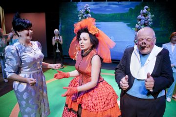 Lavalla esiintyy kolme näyttelijää värikkäissä, dramaattisissa puvuissa. Punapukuinen nainen keskellä näyttää levottomalta, ja häntä reunustavat hymyilevä violettipukuinen nainen ja peukkua ylöspäin heiluttava kalju mies, joiden takana on maalattu luontotausta.