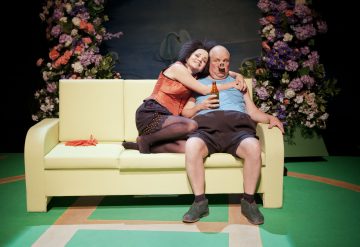 Oranssiin toppiin pukeutunut nainen halajaa siniseen toppiin pukeutunutta miestä, jolla on kädessään olutpullo, kun he istuvat keltaisella sohvalla. Molemmat ovat lavasteessa, jonka taustalla on värikkäitä kukka-asetelmia.