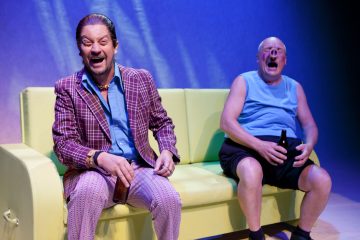 Kaksi miestä istuu keltaisella sohvalla näyttämövalaistuksen alla; toisella on kuvioitu puku ja hän nauraa, kun taas toinen, siniseen toppiin ja shortseihin pukeutunut mies nauraa myös yllättyneenä ja pitelee pulloa.