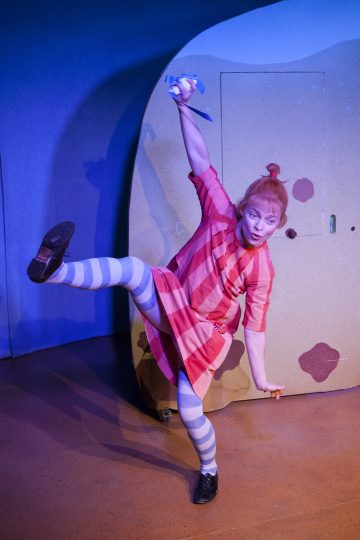 Punaraitaiseen mekkoon ja sinivalkoraidallisiin raidallisiin sukkahousuihin pukeutunut esiintyjä poseeraa lavalla leikkisästi, nostaa toisen kätensä paperiesineen kanssa ja nostaa toisen jalkansa, ja hänen takanaan on omituinen tausta.