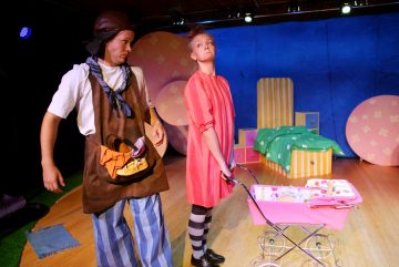 Lavalla esiintyy kaksi näyttelijää, joista toisella on ruskea esiliina, huivi ja hattu, kun taas toisella on löysä punainen mekko ja raidalliset sukkahousut, ja hän työntää vaaleanpunaisia lelukärryjä. Värikäs lavastus sisältää sänkyjä ja suuria pyöreitä muotoja.