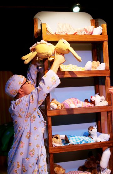 Siniseen pyjamaan pukeutunut henkilö, jolla on nallenpainatuksia, asettaa keltaisen pehmoeläimen puiseen kerrossänkyyn, joka on täynnä erilaisia pehmoleluja, kirkkaan ylävalon alla.