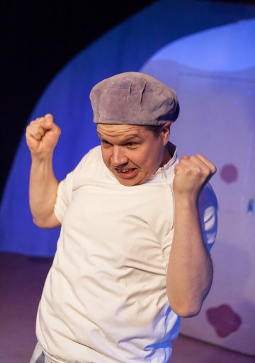 Henkilö, jolla on vaaleanvioletti lippis ja valkoinen paita, puristaa molemmat nyrkkinsä ja nostaa ne kasvojensa lähelle, vaikuttaen kiihtyneeltä tai intensiiviseltä, ja hänen takanaan on sininen ja violetti tausta.