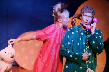 Lavalla esiintyy kaksi värikkäisiin pukuihin pukeutunutta näyttelijää; toinen vaaleanpunaisessa mekossa, jossa on pehmokissa, ja toinen vihreässä pilkkupuvussa, jossa on peruukki, ja puhuu vaaleanpunaiseen lelupuhelimeen. Taustaa koristavat pyöreät oranssit muodot.