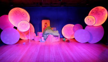 Henkilö istuu lavalla sängyssä, jota ympäröivät suuret, pyöreät, hehkuvat lavasteet, joissa on sarjakuvamaisia sian kasvoja. Tausta on tummansininen ja lattia on puinen, mikä luo omituisen, leikkisän tunnelman.