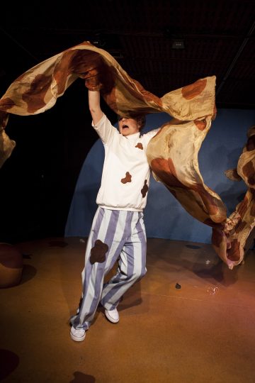 Esiintyjä, jolla on raidalliset housut ja valkoinen paita, jossa on ruskeita pilkkuja, heiluttaa suurta, ruskeapilkkuista kangasta näyttämön yläpuolella luoden dynaamisen ja energisen liikkeen tummaa taustaa vasten.