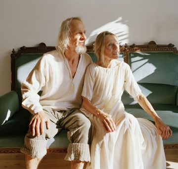 Iäkäs pariskunta istuu vintage-vihreällä sohvalla kirkkaassa auringonvalossa. Molemmilla on kevyet, väljät vaatteet ja he katsovat rauhallisin ilmein kauas. Kasvin varjot lankeavat heidän takanaan olevalle seinälle.