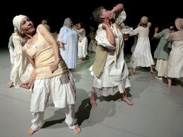 Ryhmä paljasjalkaisia ihmisiä, joilla on löysät, valkoiset, aikakauden tyyliin sopivat vaatteet, esittää näyttämöllä ilmeikkäitä tanssiliikkeitä, osa etualalla poseeraa tarmokkaasti ja osa on vuorovaikutuksessa taustalla.