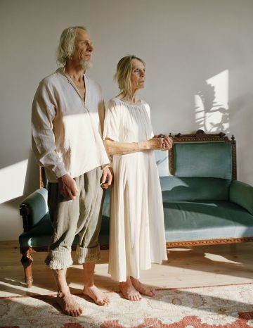 Iäkäs mies ja nainen seisovat paljain jaloin matolla, pitävät toisiaan kädestä kiinni ja katsovat mietteliäästi kaukaisuuteen. Heillä on väljät, vaaleat vaatteet, ja he seisovat auringonvalossa olevan huoneen vihreän vanhan sohvan edessä.