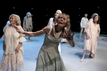 Ryhmä vaaleisiin, väljiin vaatteisiin pukeutuneita vanhempia aikuisia seisoo lavalla paljain jaloin, jotkut venyttelevät käsiään tai katsovat ylöspäin intensiiviset, ilmeikkäät kasvot ikään kuin esittäisivät tunnekohtausta dramaattisessa valaistuksessa.