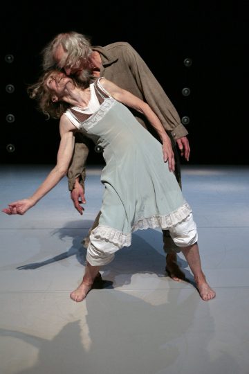 Vanhempi mies ja nainen esittävät nykytanssia lavalla. Nainen nojaa sirosti taaksepäin, kädet ojennettuina, kun taas mies seisoo lähellä hänen takanaan. Molemmat ovat paljain jaloin ja pukeutuneet väljiin, rentoihin vaatteisiin.