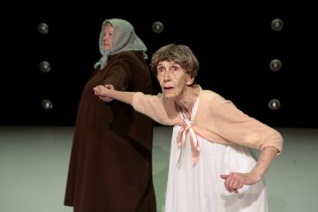 Kaksi iäkästä naista seisoo kädestä pitäen. Toisella on ruskea takki ja päähuivi, ja hän on vasemmalle päin, kun taas toinen, jolla on valkoinen mekko ja vaaleanpunainen villatakki, on kasvot eteenpäin, käsi ojennettuna ja huolestuneen näköisenä. Taustalla on tumma pyöreä valo.