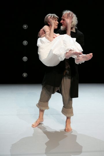 Harmaahiuksinen ja parrakas vanhempi mies seisoo lavalla paljain jaloin ja kantaa hellästi valkoiseen mekkoon pukeutunutta vanhempaa naista. He tuijottavat toisiaan tummalla, pyöreillä valoilla koristeltua taustaa vasten.