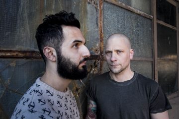 Kaksi miestä seisoo ränsistyneen, rikkinäisen ikkunan edessä. Toisella on täysi parta ja tummat hiukset, ja hän katsoo sivuttain; toinen on kalju ja hänellä on vaalea harja, ja hän katsoo eteenpäin. Molemmilla on vakavat ilmeet.