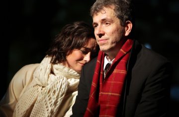 Valkoiseen villapaitaan ja huiviin pukeutunut nainen laskee päänsä mustaan takkiin ja punaiseen huiviin pukeutuneen miehen olkapäälle. Mies näyttää mietteliäältä, kun taas nainen vaikuttaa lohdutetulta, molemmat istuvat tiiviisti vierekkäin.