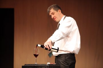 Valkoiseen paitaan ja solmioon pukeutunut mies kaataa punaviiniä pullosta pöydällä olevaan lasiin, ja hänen takanaan on neutraali ruskea tausta.