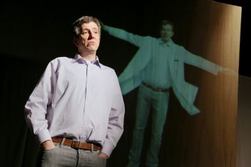 Vaalean violettiin paitaan pukeutunut mies seisoo kädet taskuissa ja näyttää vakavalta, kun hänen isompi varjonsa tai holografinen heijastuksensa, jolla on ojennetut kädet, ilmestyy seinälle hänen takanaan.