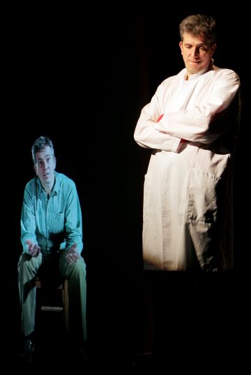 Valkoiseen laboratoriotakkiin pukeutunut mies seisoo kädet ristissä ja katsoo istuvaa miestä, joka näyttää aavemaiselta tai hologrammilta tummaa taustaa vasten.