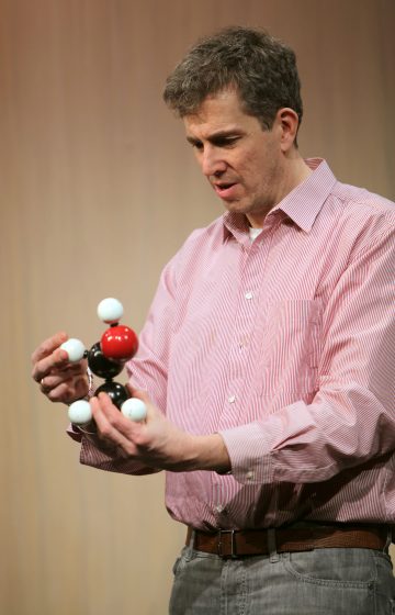 Vaaleanpunaiseen raidalliseen paitaan pukeutunut mies tutkii ja tarkastelee molekyylimallia, joka koostuu sauvoilla yhdistetyistä mustista, valkoisista ja punaisista palloista. Hän vaikuttaa keskittyneeltä ja seisoo tavallista vaaleaa taustaa vasten.