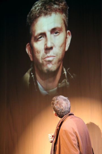 Ruskeaan takkiin pukeutunut mies katsoo seinälle heijastettua suurta kuvaa toisen miehen kasvoista, mikä luo dramaattisen ja intensiivisen tunnelman.