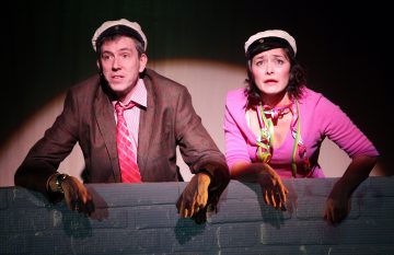 Kaksi näyttelijää, joilla on hattu ja ilmeikkäät kasvot, nojaavat näyttämöllä matalan seinän yli, jota valonheitin valaisee. Miehellä on ruudullinen takki ja punainen solmio; naisella on vaaleanpunainen villatakki, jonka kaulassa on värikkäitä nauhoja.