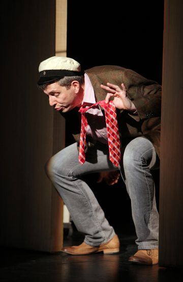 Mies, jolla on kuviollinen bleiseri, punaruutuinen solmio ja merimieshattu, kyyristyy oviaukossa ja tekee kädellään OK-eleen nojatessaan eteenpäin leikkisällä ilmeellä lavalle.